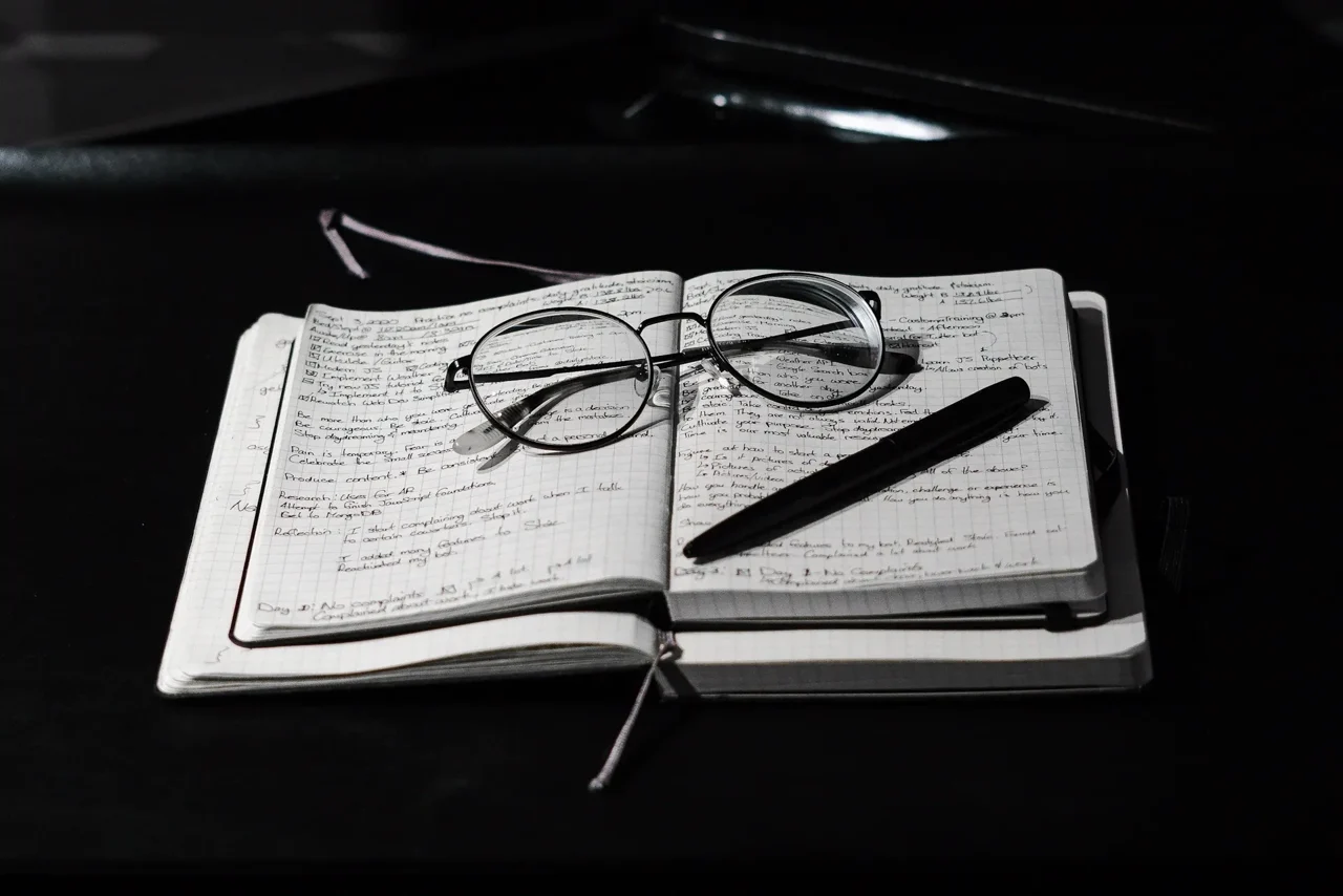 A notebook of handwritten notes, glasses and a pen on a desk.
