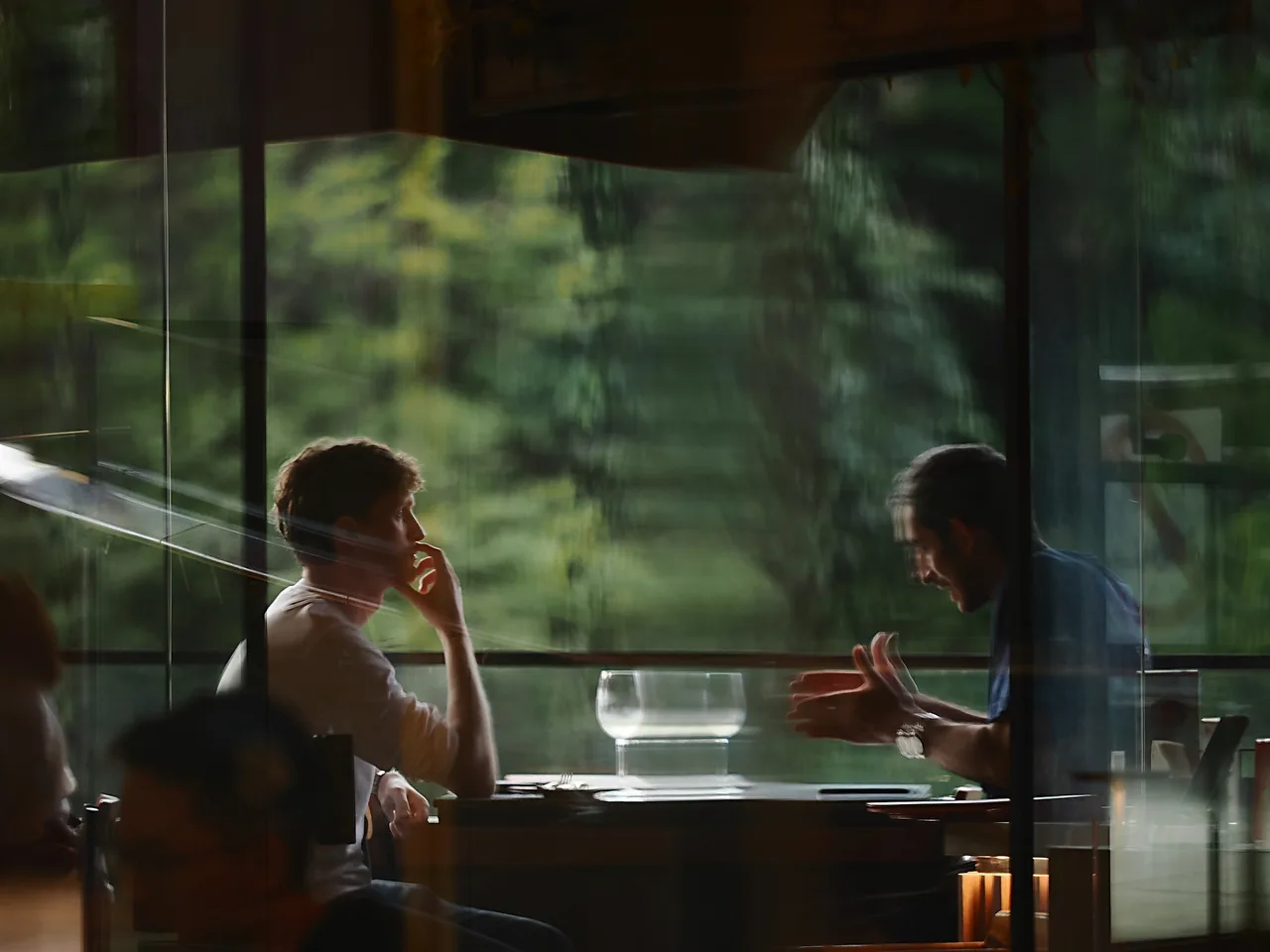 Two people in conversation at a table, seen through a window.